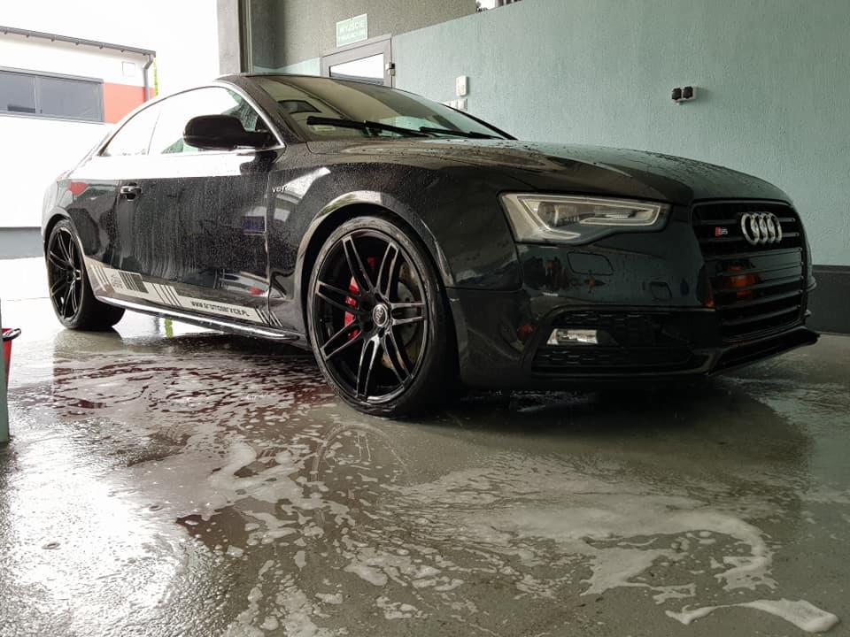 Czarny samochód Audi S5 po umyciu, z kroplami wody na karoserii i mokrą posadzką pokrytą pianą, widoczne czerwone zaciski hamulcowe i czarne felgi.