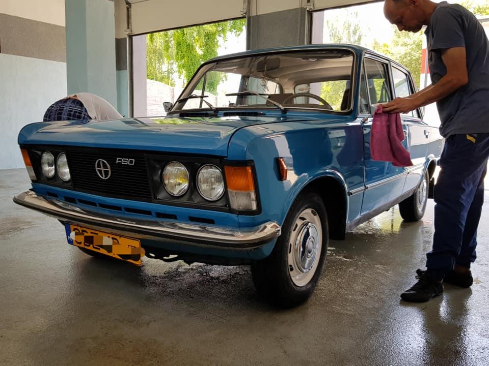 Mężczyzna poleruje turkusowy samochód FSO 125p szmatką w myjni, widoczne detale karoserii i reflektorów.