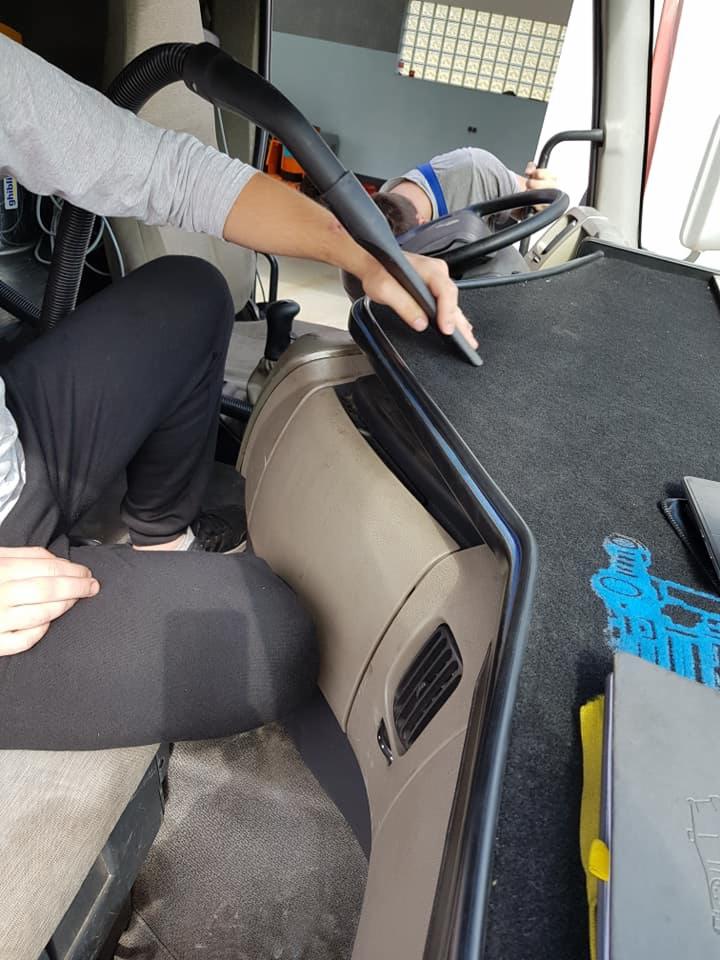 Czyszczenie wnętrza kabiny pojazdu za pomocą odkurzacza przemysłowego, widoczne elementy deski rozdzielczej i dywanika podłogowego z niebieskim wzorem.