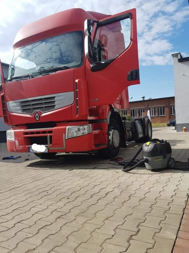 Czerwona kabina ciężarówki z otwartymi drzwiami, odkurzacz przemysłowy Kärcher na kostce brukowej, prawdopodobnie podczas czyszczenia wnętrza pojazdu.