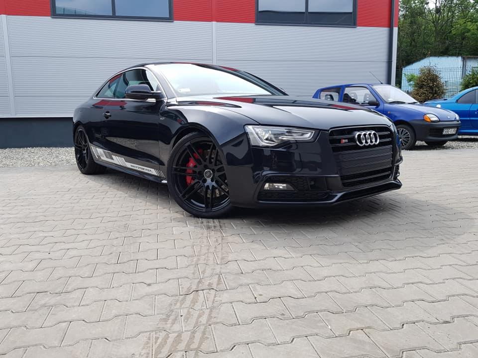 Błyszczący, czarny samochód Audi S5 Coupe z czerwonymi zaciskami hamulcowymi na tle szarego budynku z czerwonymi akcentami i kostki brukowej.