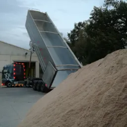 Ciężarówka MAN z podniesioną naczepą wyładowuje piasek lub żwir na placu budowy, widoczny budynek magazynowy w tle.