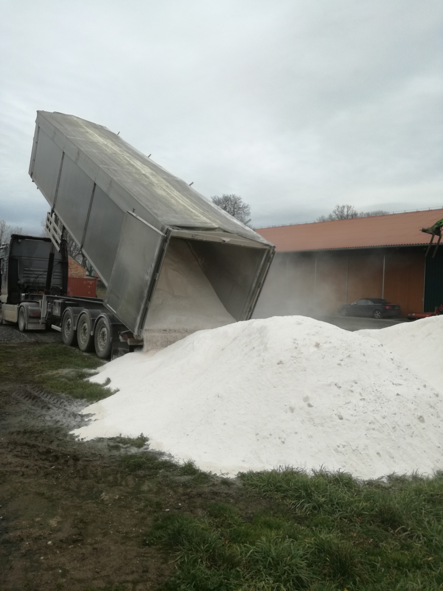 Ciężarówka z naczepą w Jurowcach wysypuje biały materiał sypki, prawdopodobnie wapno, tworząc dużą pryzmę na ziemi, z widocznymi śladami opon na błotnistym podłożu.