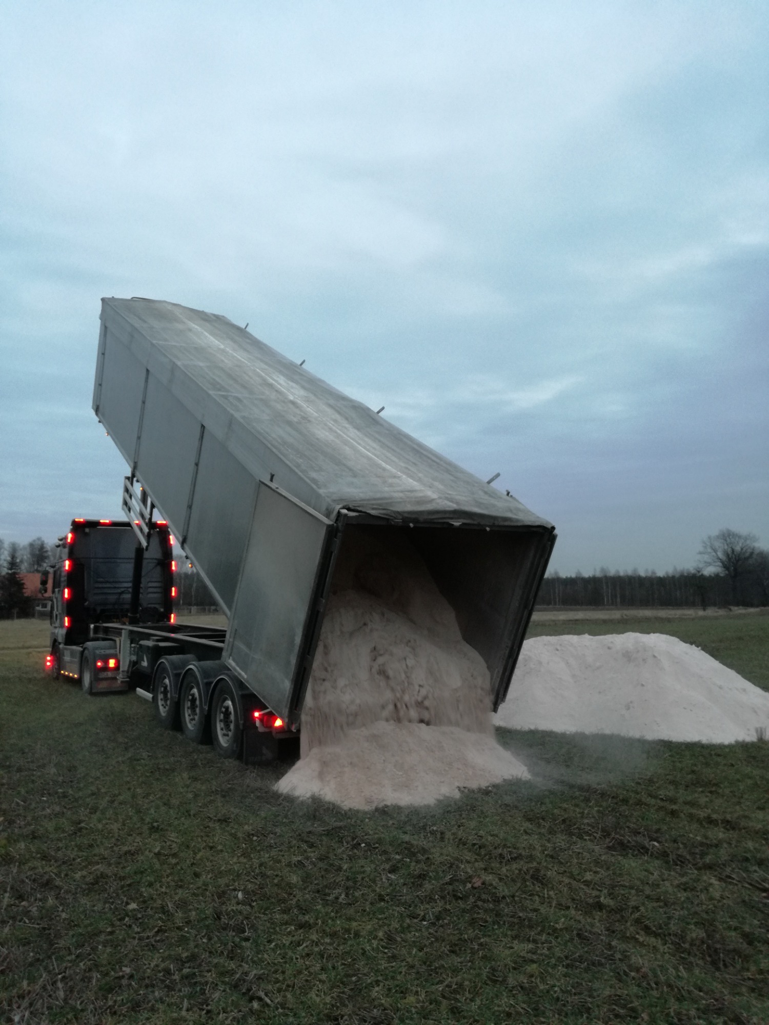 Ciężarówka z naczepą wywrotką, w trakcie rozładunku jasnego materiału sypkiego na zielone pole, widoczne światła pozycyjne i tylne pojazdu, ponure niebo w tle.