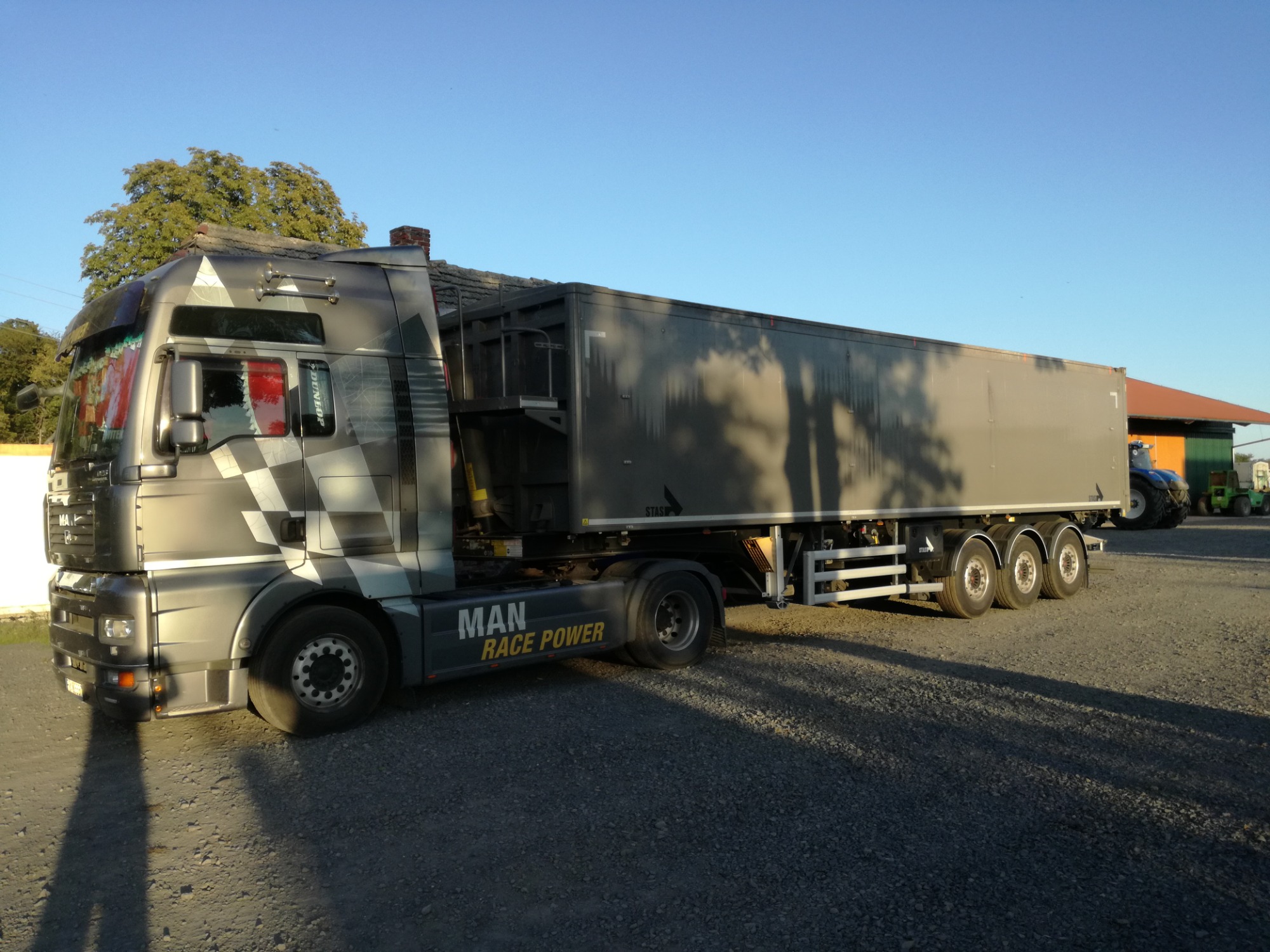 Szaro-czarny ciągnik siodłowy MAN z naczepą, ozdobiony motywem szachownicy na kabinie i napisem 'MAN RACE POWER' na błotniku, zaparkowany na żwirowym placu w słoneczny dzień.