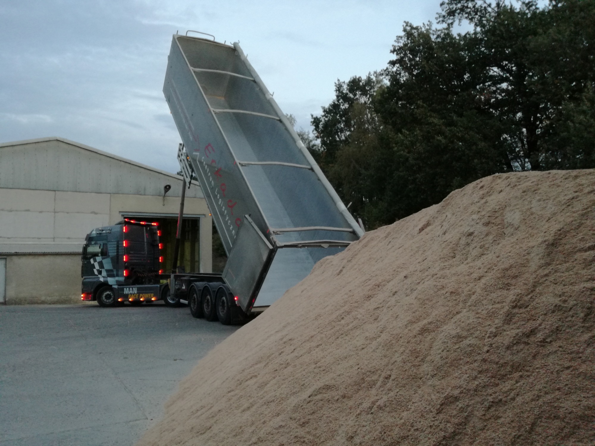 Ciężarówka MAN z podniesioną naczepą wyładowuje piasek lub żwir na placu budowy, widoczny budynek magazynowy w tle.