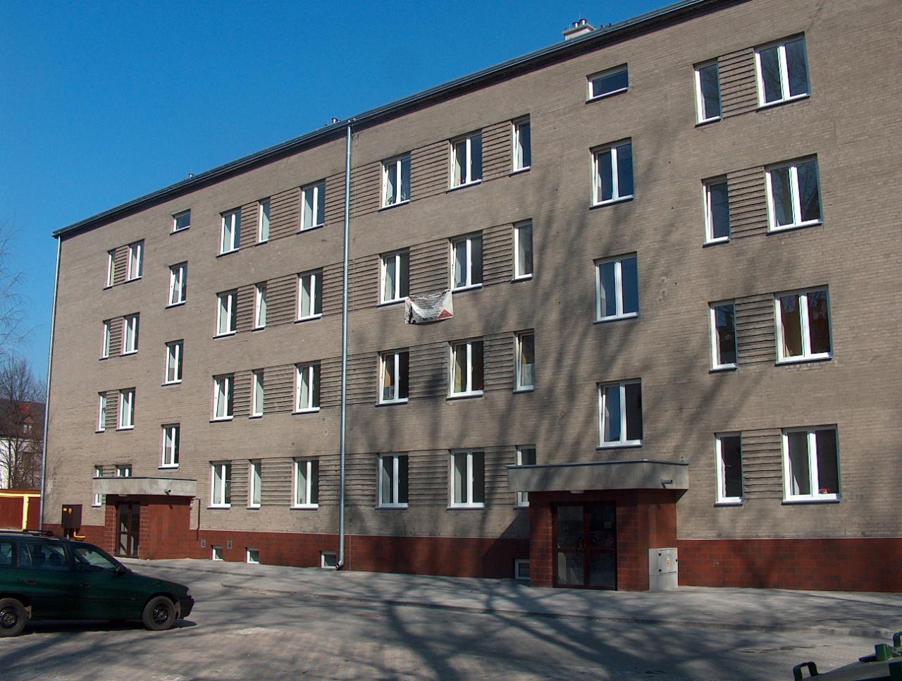 Wielorodzinny budynek z szarej cegły z widoczną instalacją odgromową w postaci pionowej rury na fasadzie, przyłącze elektryczne do budynku, oraz skrzynka elektryczna przy wejściu.