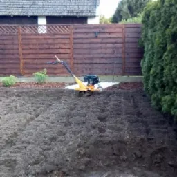 Żółty glebogryzarek spalinowy na przygotowanym do siewu, spulchnionym gruncie w ogrodzie, z drewnianym płotem i żywopłotem w tle.