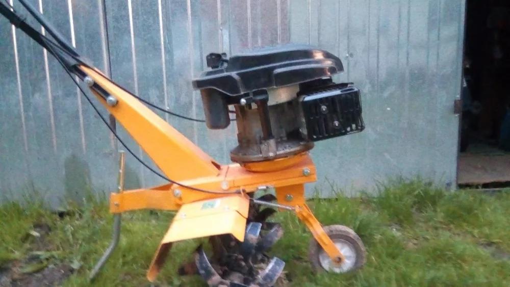 Pomarańczowy glebogryzarkę spalinowa, stojąca na trawie przed blaszanym garażem, widoczne elementy robocze z resztkami ziemi.