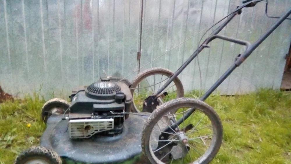 Używana kosiarka spalinowa, z kołami rowerowymi, stojąca na trawie przed blaszakiem.