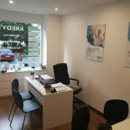 Wnętrze biura z biurkiem, krzesłami, laptopem i kwiatami. Na ścianie plakaty reklamowe z ofertą finansową. W oknie widoczny napis 'Kredyty' i czerwony samochód.