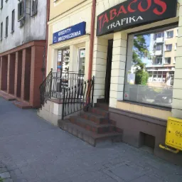 Fasada budynku z biurem kredytów i ubezpieczeń obok kiosku Trafika, widoczne schody wejściowe z czarną balustradą i niebieski szyld 'Kredyty Ubezpieczenia'.