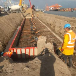 Zbrojenie fundamentów w wykopie na terenie portu, widoczny pracownik w odblaskowej kamizelce i kasku, betonowozy w tle.