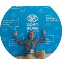 Mężczyzna z identyfikatorem na tle niebieskiej ściany z logo 'open eyes economy summit' i hasłami w dymkach, uśmiechnięty, z rozłożonymi rękami.