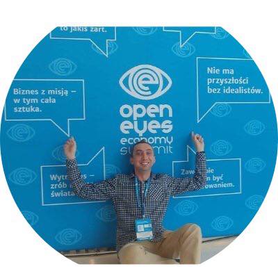 Mężczyzna z identyfikatorem na tle niebieskiej ściany z logo 'open eyes economy summit' i hasłami w dymkach, uśmiechnięty, z rozłożonymi rękami.