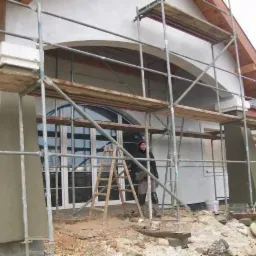 Prace przygotowawcze do wykończenia elewacji w Wadowicach: kolumna z nałożonym tynkiem w kolorze piaskowym, rusztowanie i drabina przed budynkiem z dużymi oknami.