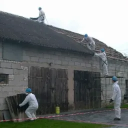 Demontaż eternitu z dachu budynku gospodarczego przez ekipę w kombinezonach ochronnych, widoczny żółty znak ostrzegawczy o zagrożeniu azbestem.