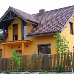 Żółty dom jednorodzinny z brązową dachówką i drewnianym ogrodzeniem, widok z zewnątrz, z elementami dekoracyjnymi z ciemnego drewna w szczycie dachu i na balkonie.