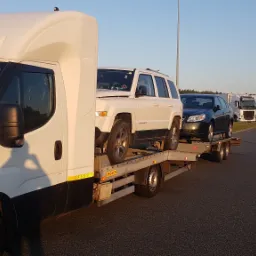 Laweta z dwoma samochodami osobowymi (biały Jeep Patriot i ciemny Saab) na parkingu, w tle inne lawety i stacja benzynowa.
