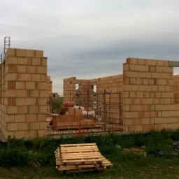 Wznoszenie ścian z bloczków ceramicznych na budowie domu jednorodzinnego, widoczne zbrojenia i paleta z drewnianymi elementami na pierwszym planie, pochmurne niebo w tle.