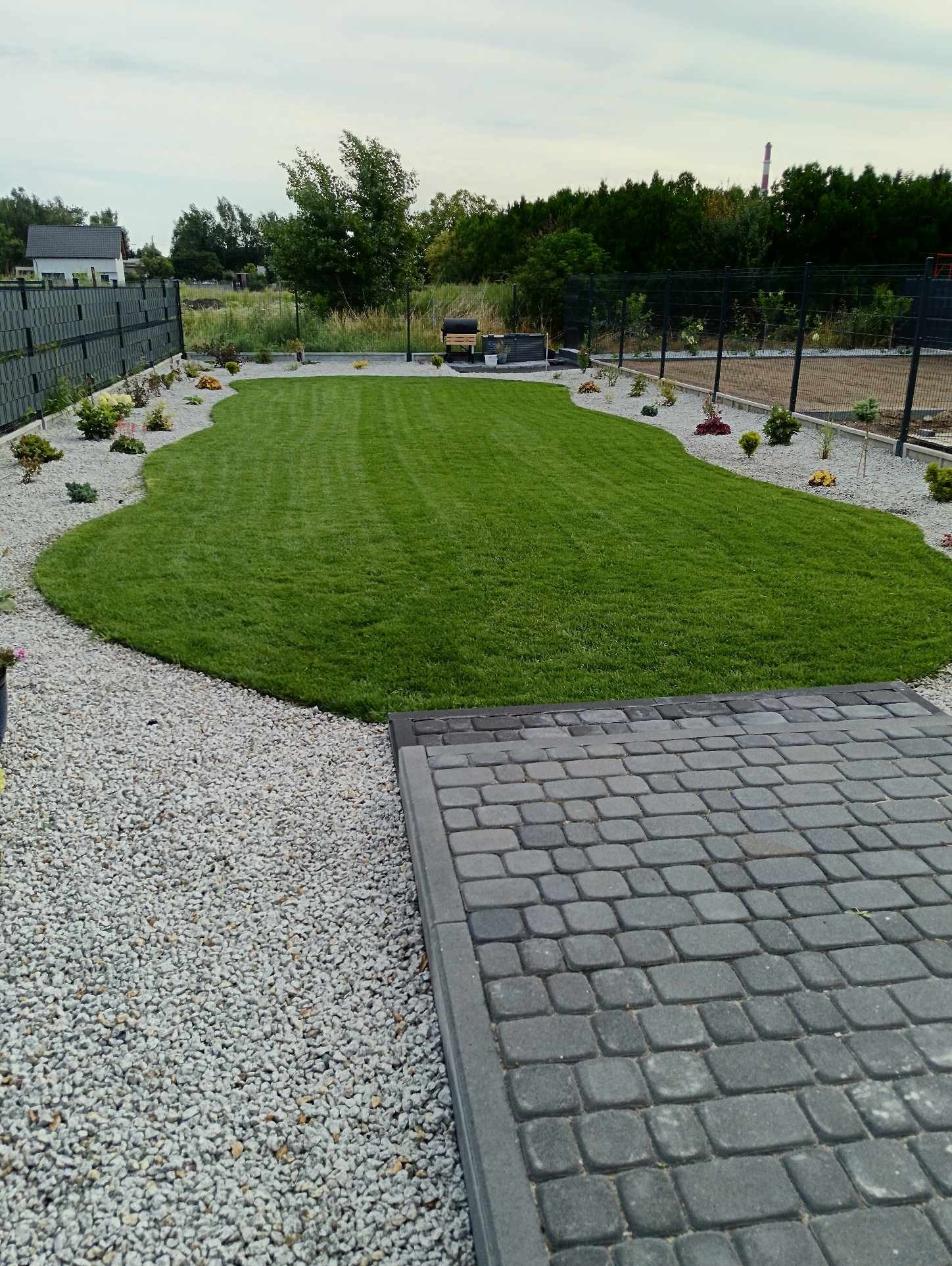 Zadbany trawnik o nieregularnym kształcie otoczony białym gresem i rabatami z niskimi roślinami, z widoczną szarą kostką brukową i ogrodzeniem panelowym w tle.