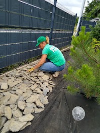 Kobieta w zielonej koszulce i czapce układa kamienie na czarnej macie wzdłuż ogrodzenia. Obok roślina iglasta i szara kula dekoracyjna, etap prac ogrodowych.