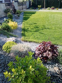 Kaskada roślinności oddzielona szarym żwirem od trawnika. Krzewy o różnych barwach i fakturach tworzą estetyczne przejście. W tle nowoczesne ogrodzenie panelowe.