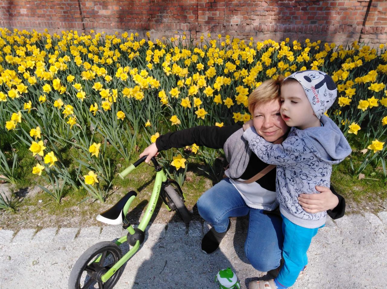 Kobieta w dżinsach i chłopiec w bluzie przytulają się na tle kwitnących żółtych narcyzów i ceglanego muru. Obok leży zielony rowerek biegowy Kinderkraft.