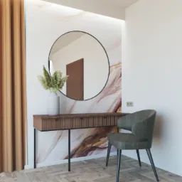 Nowoczesna toaletka z ryflowanym frontem, okrągłe lustro z czarną ramą, dekoracyjny wazon z suszonymi trawami i szare krzesło na tle ściany z marmurowym wzorem.