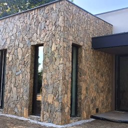 Usługi Kamieniarskie Bartłomiej Lisak - Elewacja budynku wykończona kamieniem naturalnym w odcieniach brązu i szarości, z widocznymi oknami w czarnych ramach i fragmentem tarasu z drewnianą podłogą. W tle drzewa i częściowo widoczny...