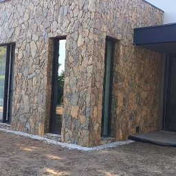 Usługi Kamieniarskie Bartłomiej Lisak - Elewacja budynku wyłożona nieregularnym kamieniem w odcieniach brązu i szarości, z widocznymi oknami i fragmentem tarasu z desek kompozytowych; teren wokół budynku jeszcze w trakcie prac...