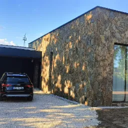 Nowoczesny dom jednorodzinny z elewacją z kamienia naturalnego, czarny samochód osobowy na żwirowym podjeździe, błękitne niebo.