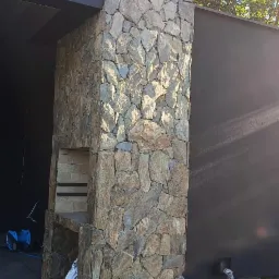 Murowany grill z kamienia naturalnego, z paleniskiem z cegły szamotowej, na betonowym tarasie, z widocznym workiem cementu i niebieskim wężem ogrodowym.