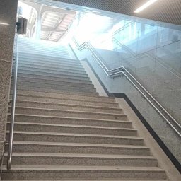 Usługi Kamieniarskie Bartłomiej Lisak - Długie kamienne schody w budynku z metalowymi poręczami i perforowanym sufitem, prowadzące do jasnego wyjścia.
