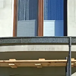 Montaż ciemnego, nakrapianego parapetu zewnętrznego pod oknem, widoczne rusztowanie i surowe wykończenie elewacji.