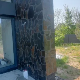 Fragment elewacji budynku z okładziną z nieregularnych, ciemnych kamieni, widoczne narzędzia i materiały budowlane, otoczenie z zielenią.