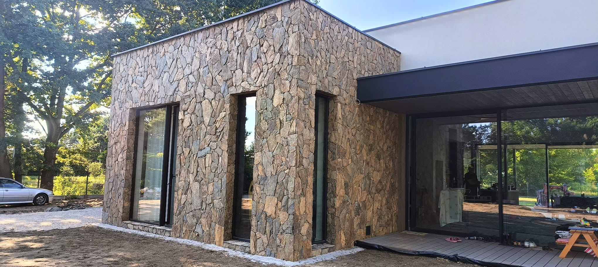 Elewacja budynku wykończona kamieniem naturalnym w odcieniach brązu i szarości, z widocznymi oknami w czarnych ramach i fragmentem tarasu z drewnianą podłogą. W tle drzewa i częściowo widoczny...