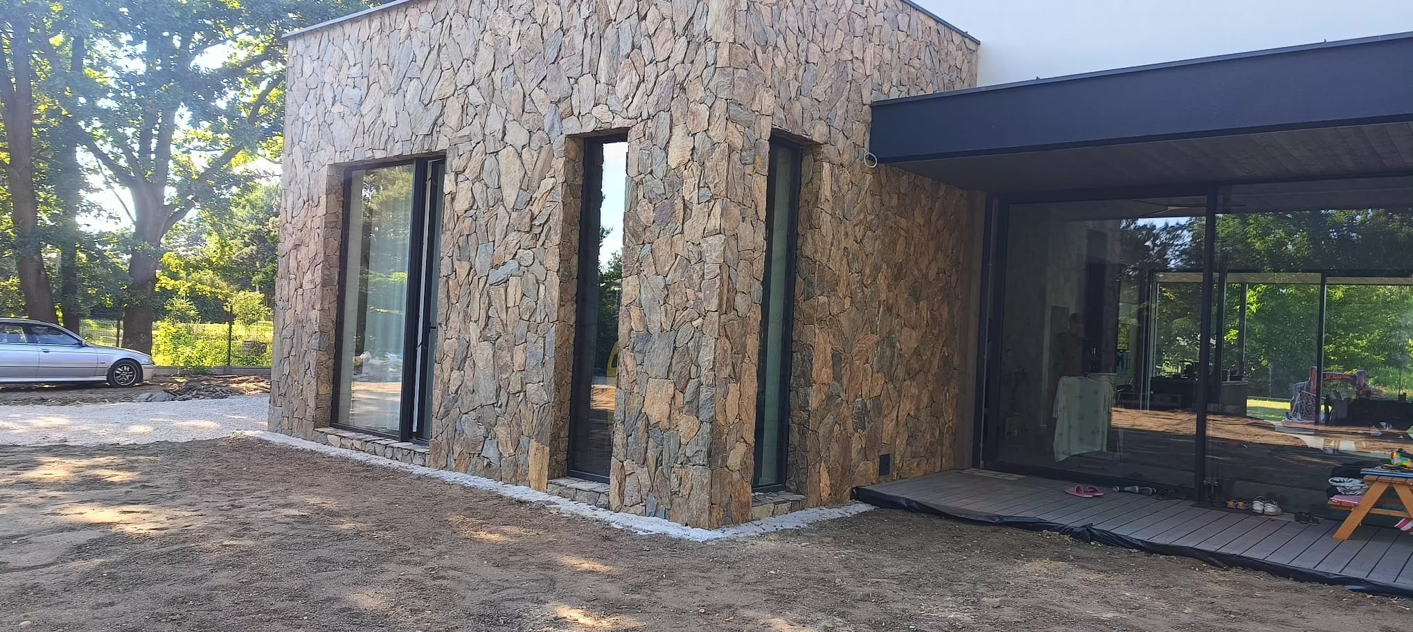 Elewacja budynku wyłożona nieregularnym kamieniem w odcieniach brązu i szarości, z widocznymi oknami i fragmentem tarasu z desek kompozytowych; teren wokół budynku jeszcze w trakcie prac...