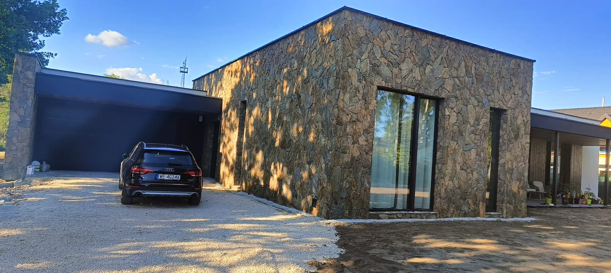 Elewacja budynku wykończona kamieniem naturalnym o nieregularnej fakturze i zróżnicowanej kolorystyce, widoczny fragment tarasu i podjazdu z czarnym Audi A4, oświetlona promieniami słońca...
