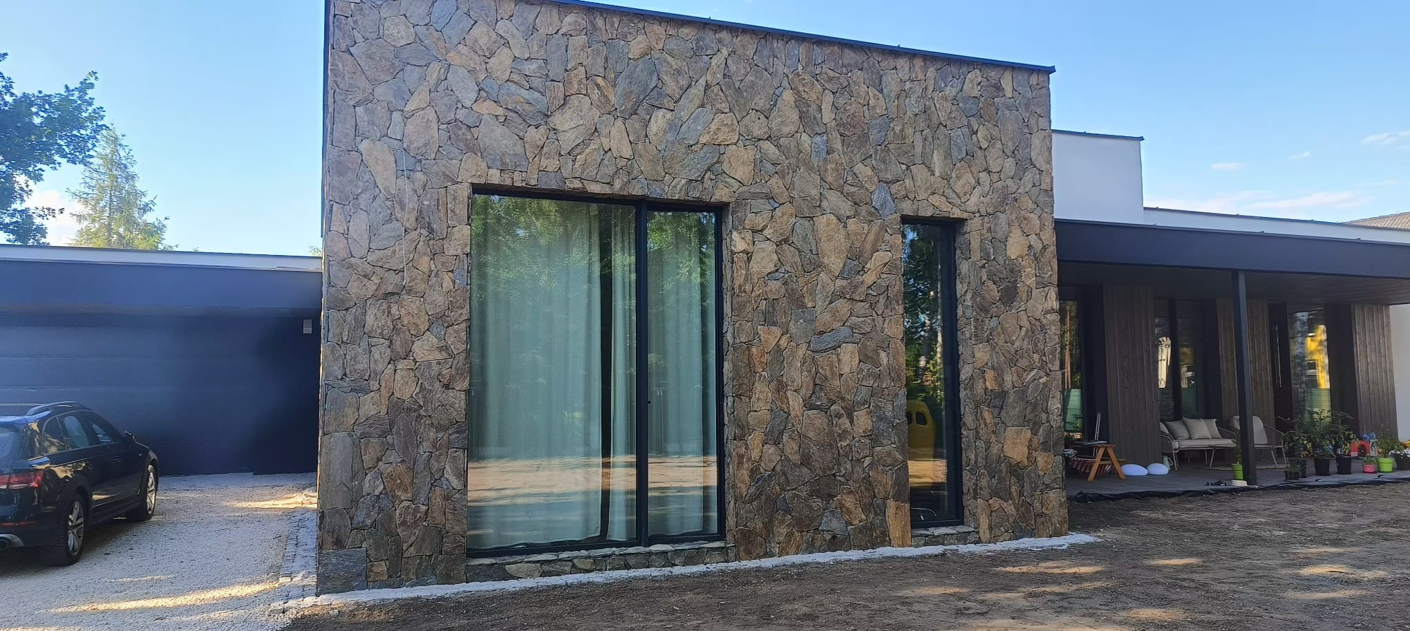 Elewacja budynku wykończona nieregularnym kamieniem naturalnym w odcieniach brązu i szarości, z dużym oknem przesuwnym i wąskim oknem stałym w czarnych ramach, widok z zewnątrz na tle błękitnego...