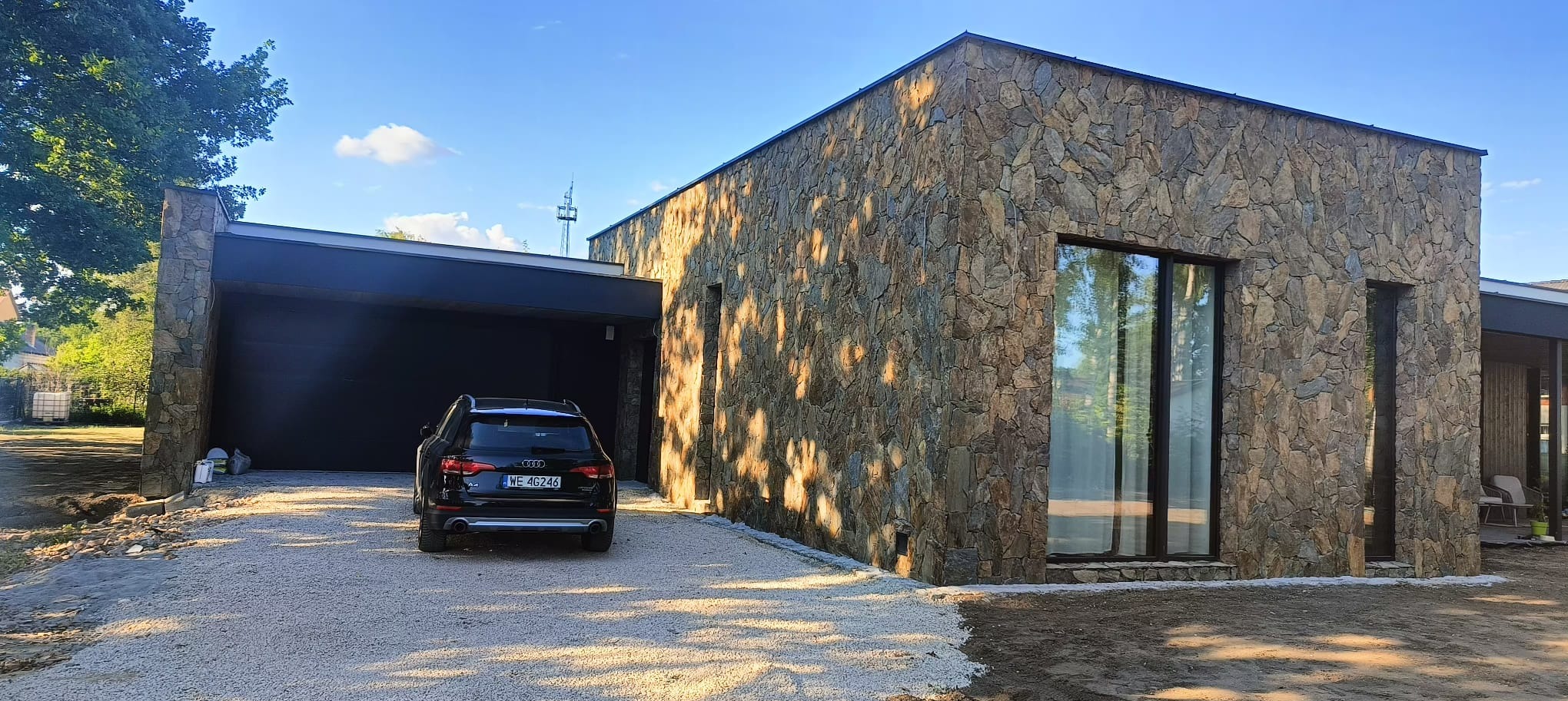 Nowoczesny dom jednorodzinny z elewacją z kamienia naturalnego, czarny samochód osobowy na żwirowym podjeździe, błękitne niebo.