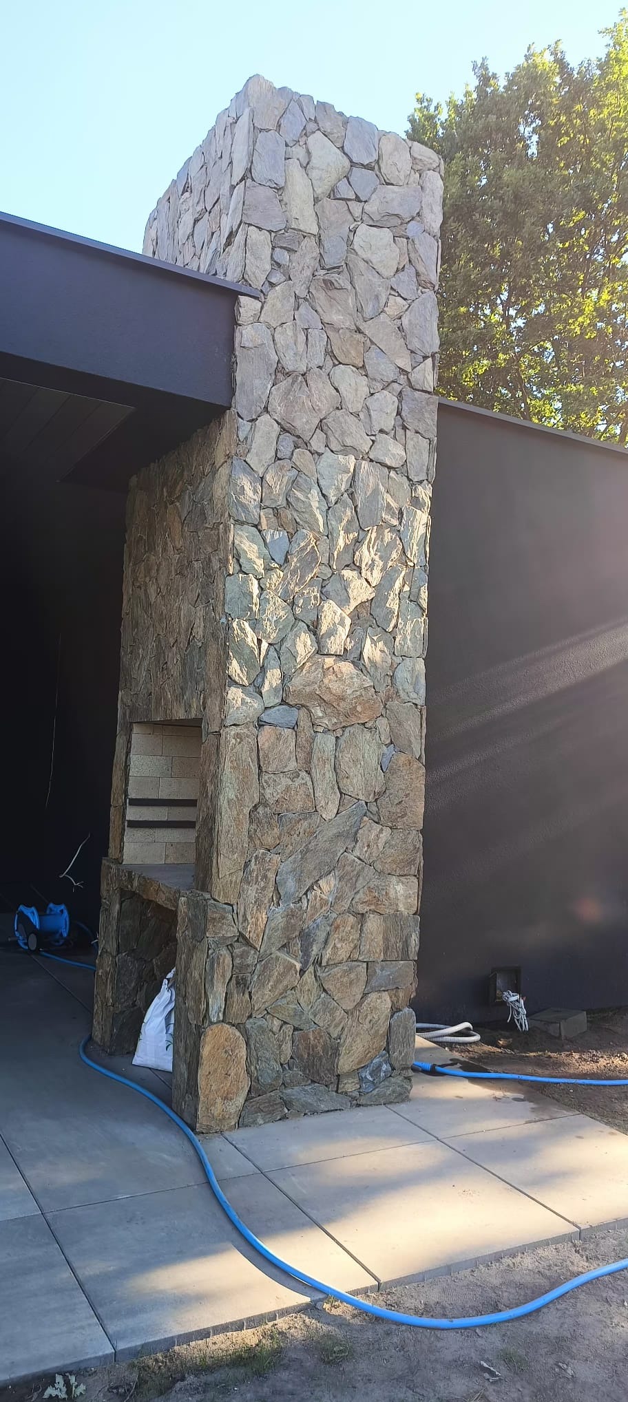 Murowany grill z kamienia naturalnego, z paleniskiem z cegły szamotowej, na betonowym tarasie, z widocznym workiem cementu i niebieskim wężem ogrodowym.