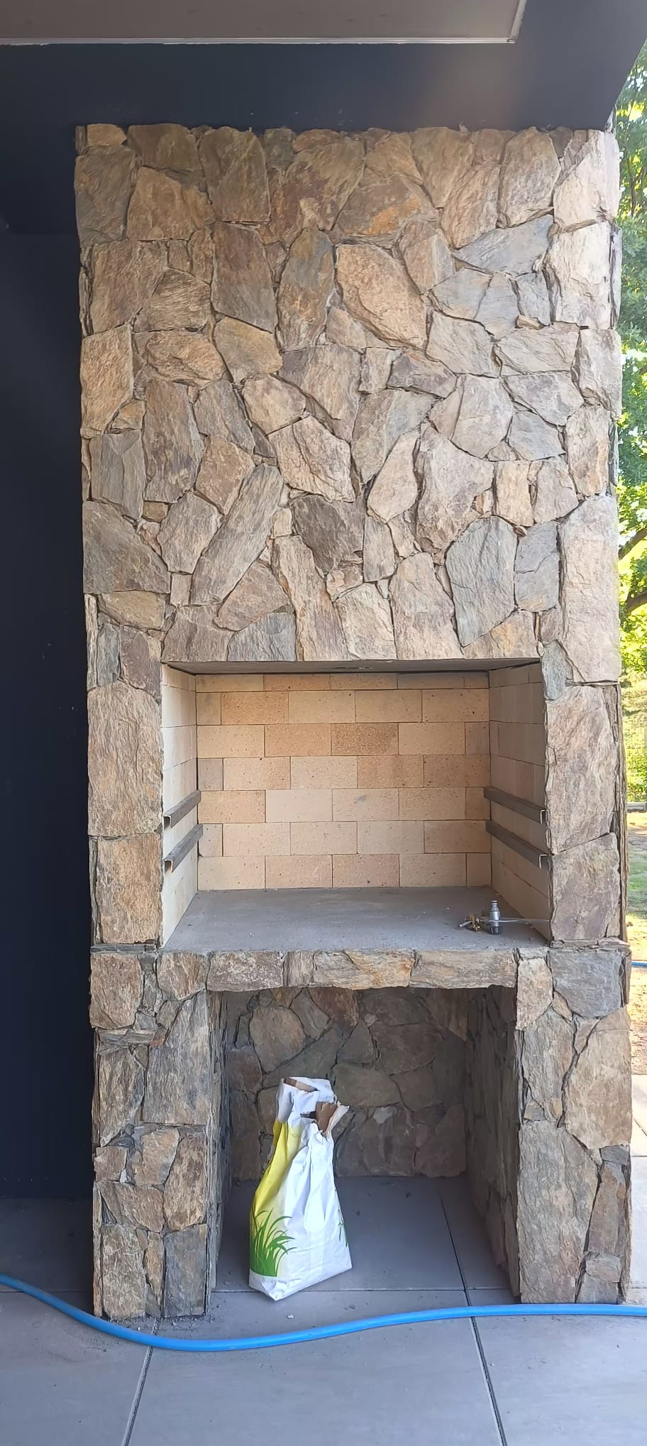 Murowany grill ogrodowy obłożony kamieniem naturalnym, z widoczną torbą z zaprawą w dolnej części.