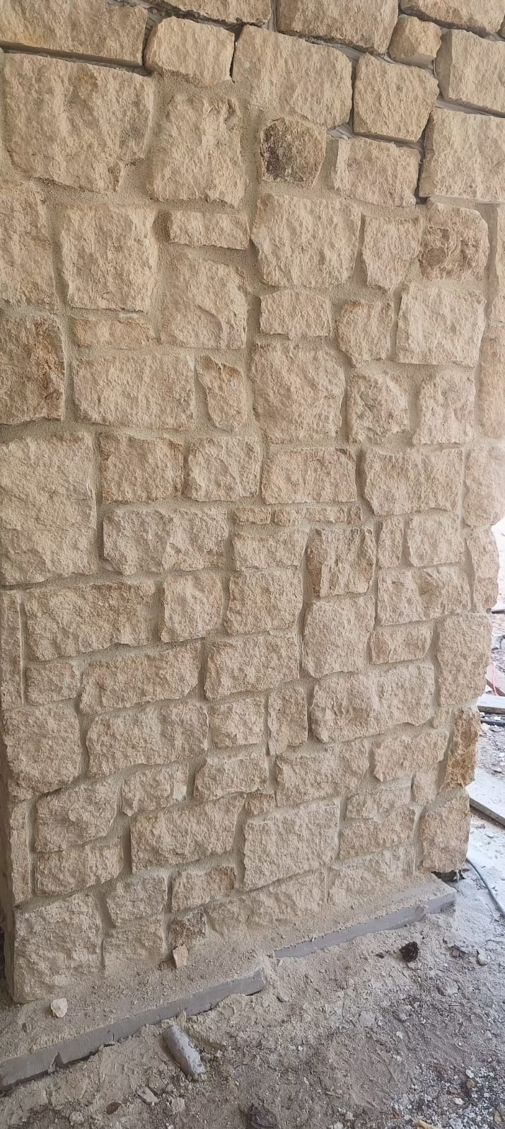 Fragment ściany wyłożonej nieregularnymi, jasnymi kamieniami z widoczną zaprawą, w trakcie budowy lub remontu.