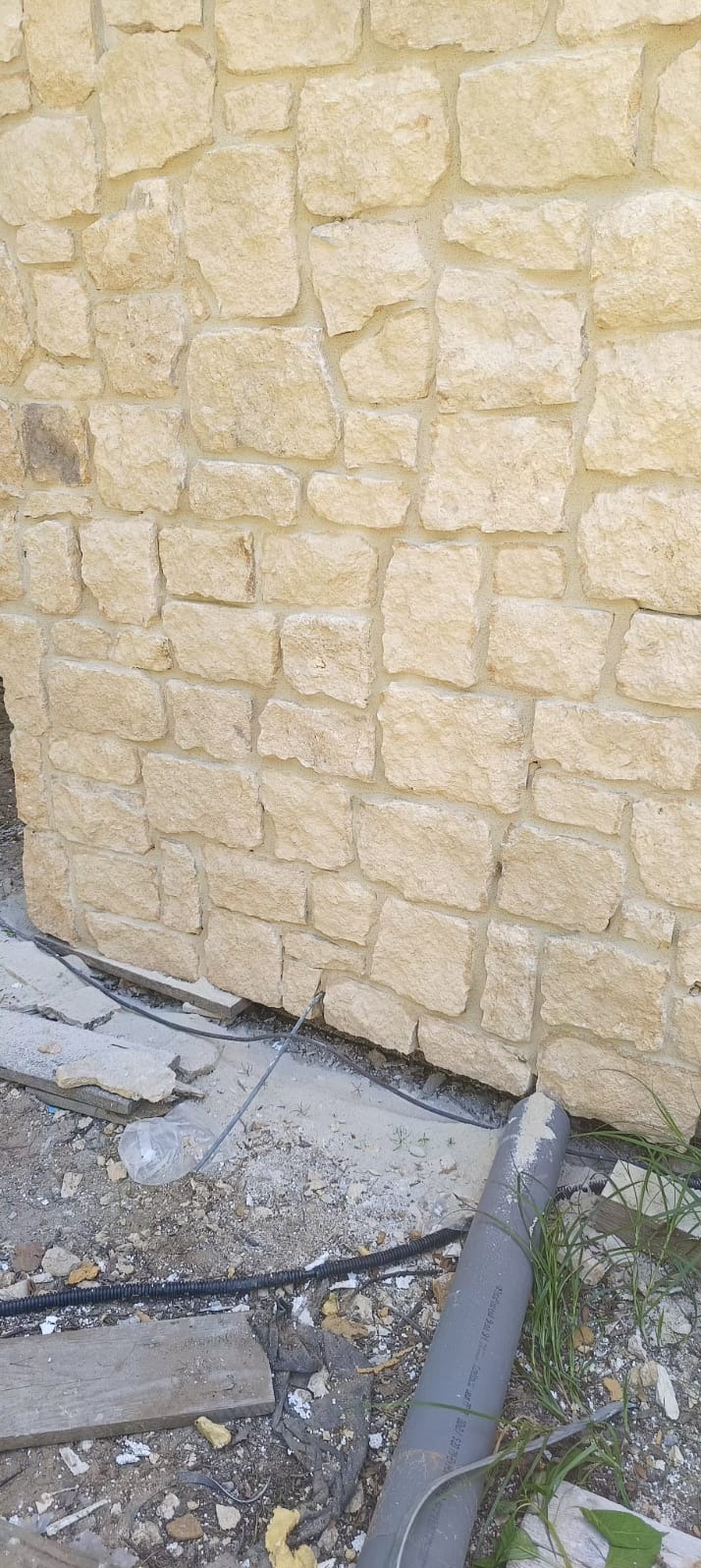Fragment ściany zewnętrznej wyłożonej nieregularnym, jasnym kamieniem, widoczne ślady zaprawy i pozostałości po budowie u podstawy ściany, w tym rura PCV.