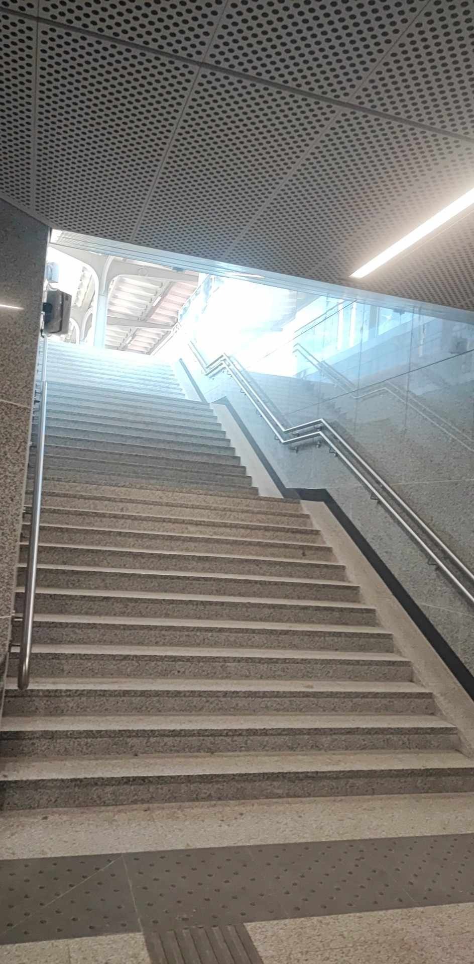 Długie kamienne schody w budynku z metalowymi poręczami i perforowanym sufitem, prowadzące do jasnego wyjścia.