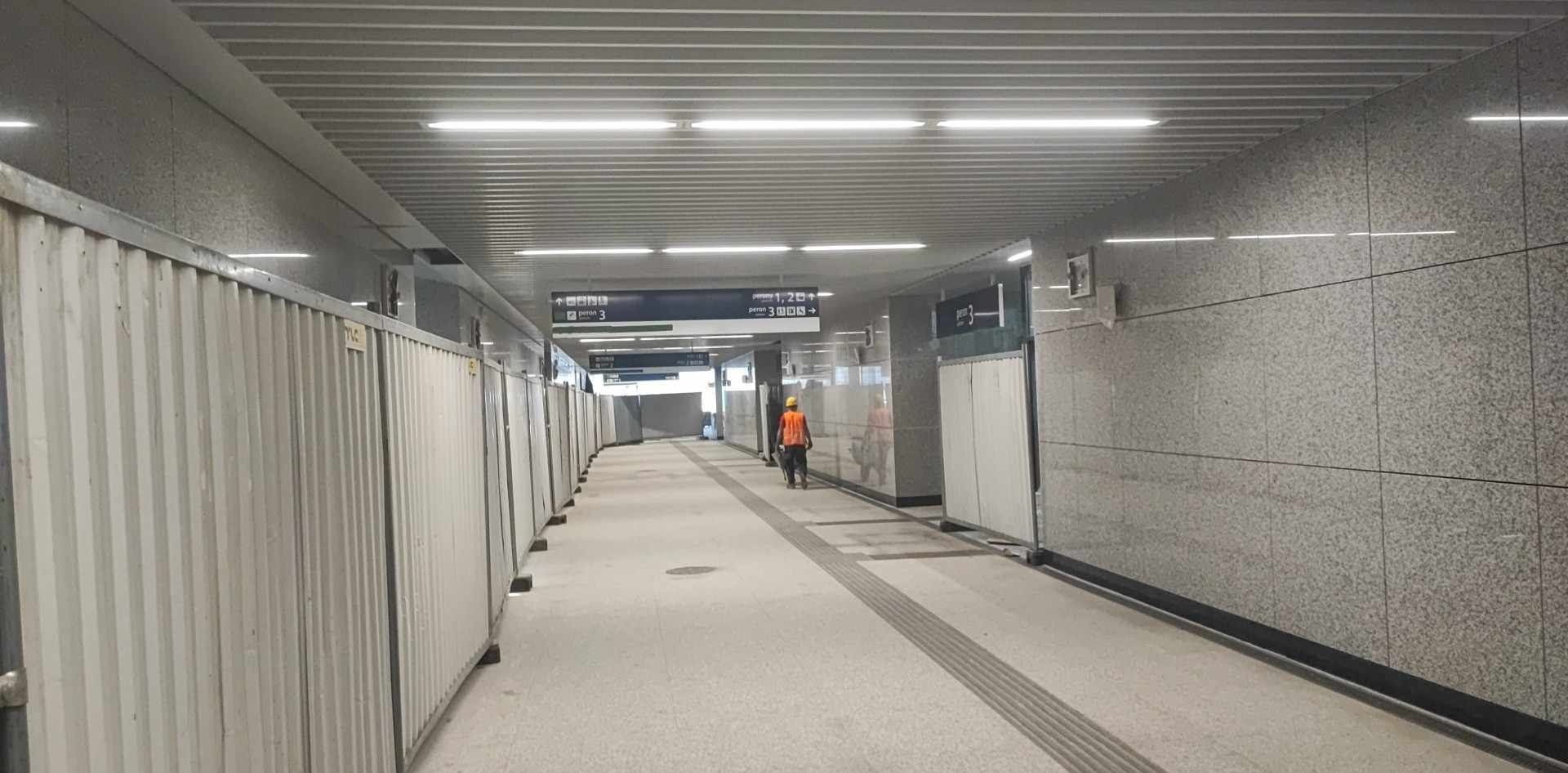 Wnętrze przejścia podziemnego w trakcie wykańczania, ściany wyłożone płytami z kamienia naturalnego, widoczne tymczasowe bariery ochronne i pracownik w stroju roboczym.