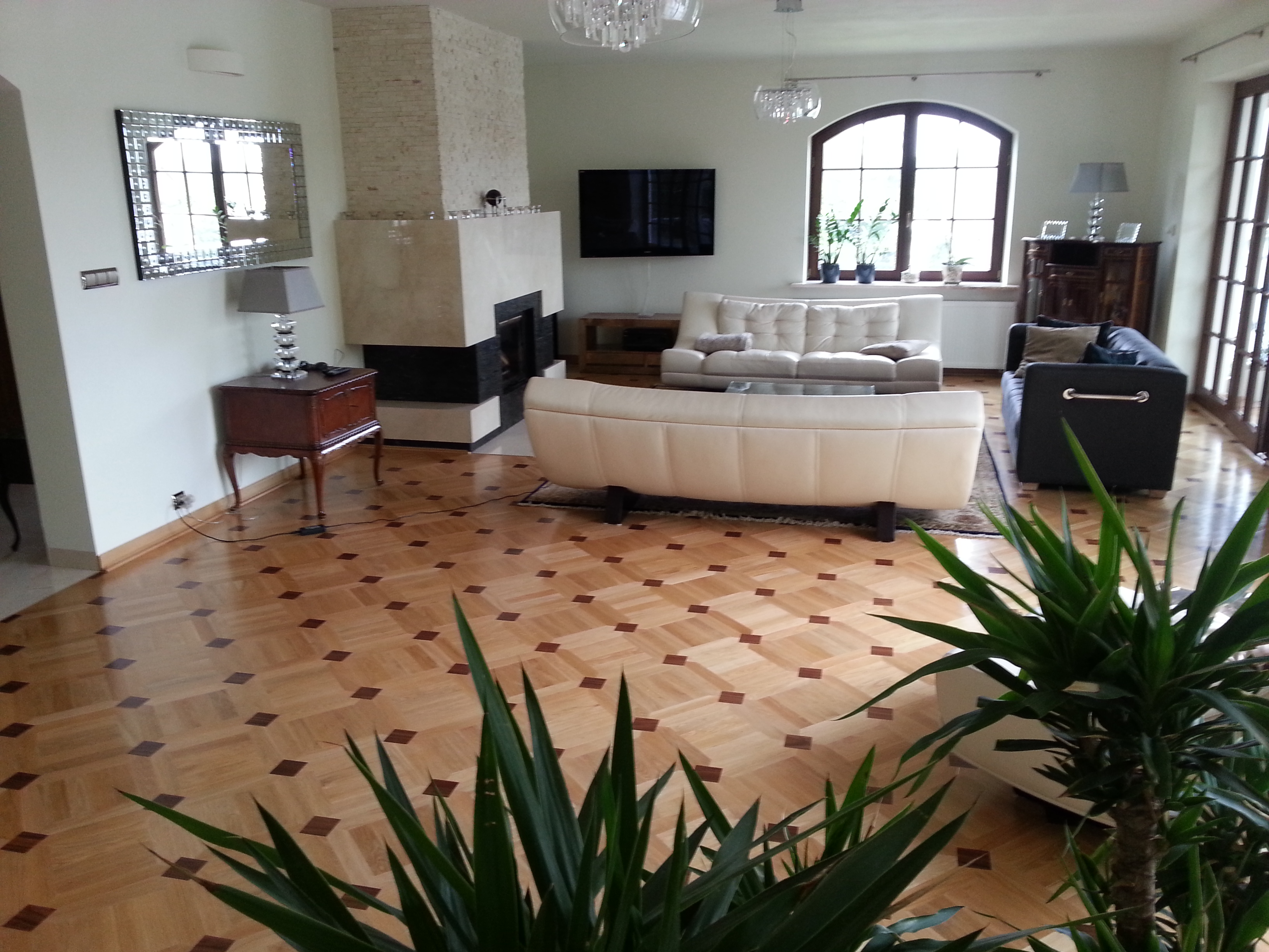 Salon z parkietem w jodełkę, kremowymi sofami i kominkiem z kamienia, widziany z perspektywy rośliny doniczkowej.