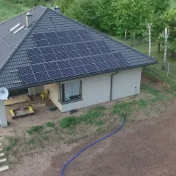 Dom jednorodzinny z czarną dachówką i panelami słonecznymi na dachu, widok z góry. Na podwórku widoczny piaskownica i ogród w trakcie przygotowania.