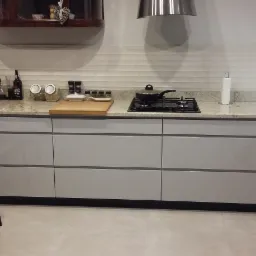 Nowoczesna, jasna kuchnia z szarymi szafkami, blatem z jasnego granitu, czarną płytą gazową z garnkiem i czarnym krzesłem o ażurowej konstrukcji.